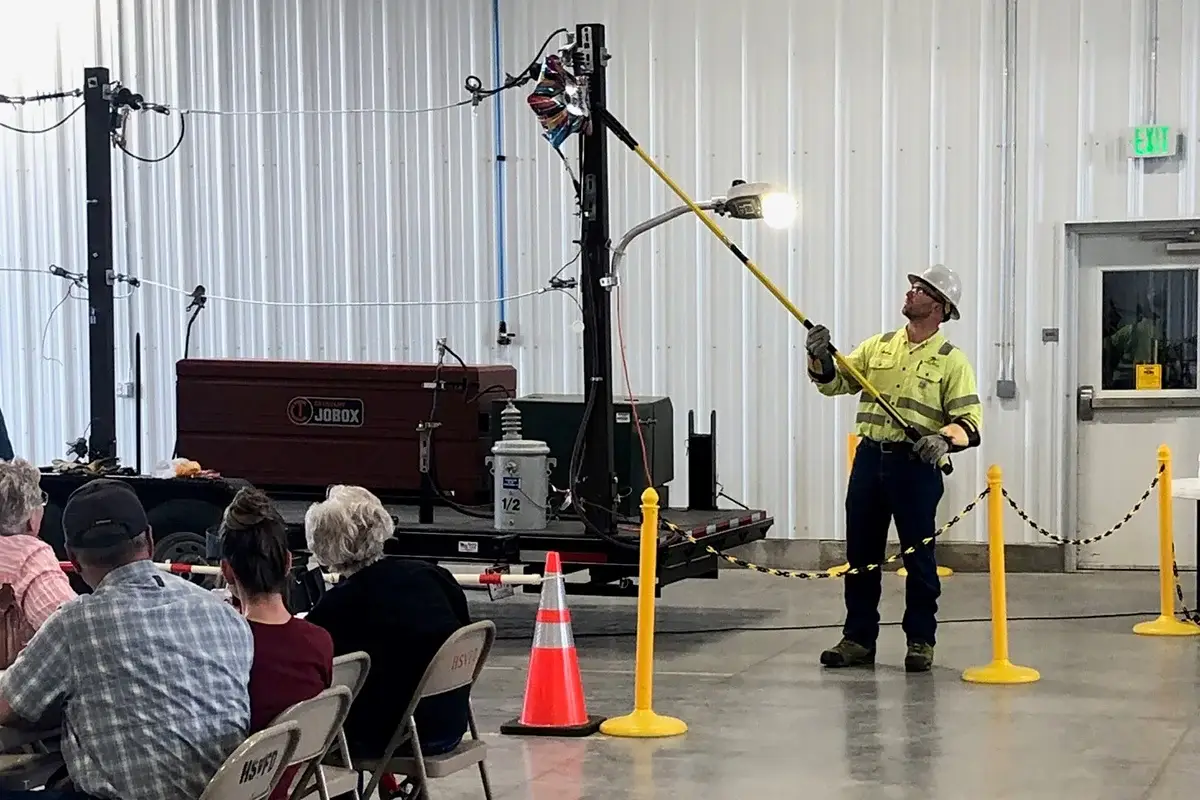 A utility worker in safety gear demonstrates electrical equipment indoors to a seated audience, using a long pole to interact with a power line model while caution cones and barriers surround the display.