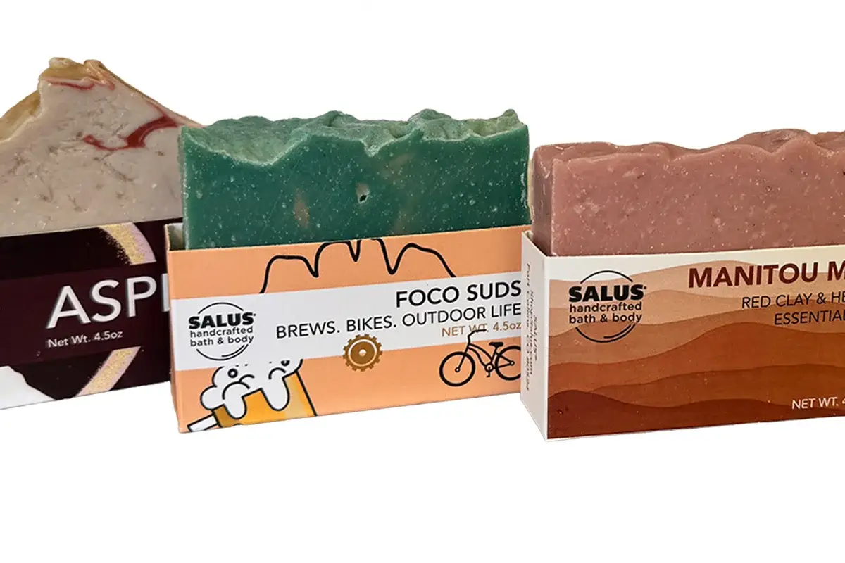 CCL Monthly Giveaway Three colorful artisan soap bars partially wrapped in labeled packaging: Aspen, Foco Suds, and Manitou Mud from Salus Bath & Body. The packaging features graphics and the soaps have textured, uneven tops.