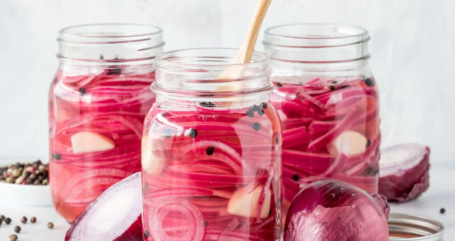 Three jars filled with sliced pickled red onions, garlic cloves, and peppercorns. A wooden spoon is in one jar. A halved onion and scattered peppercorns are on the table.