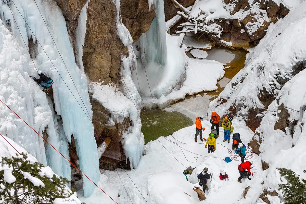 A group of people in winter gear stand near a frozen waterfall and ice formations while one person climbs the ice using ropes.