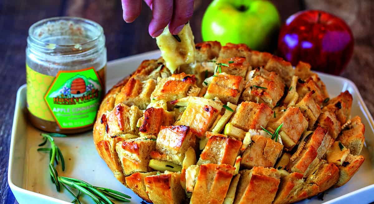 Sliced pull-apart bread served with apple slices, fresh rosemary, and apple butter on a plate. A hand is dipping a piece of bread into the apple butter.