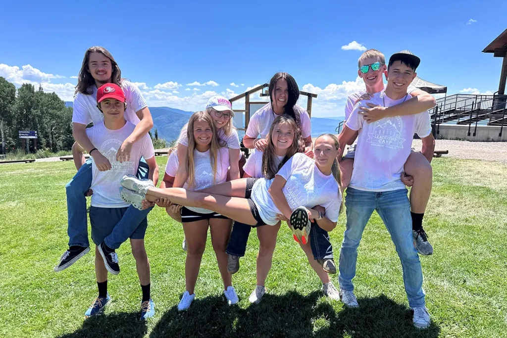 Eight teenagers pose outdoors on grass, some giving piggyback rides, all smiling and wearing matching light-colored shirts, with mountains and a blue sky in the background.