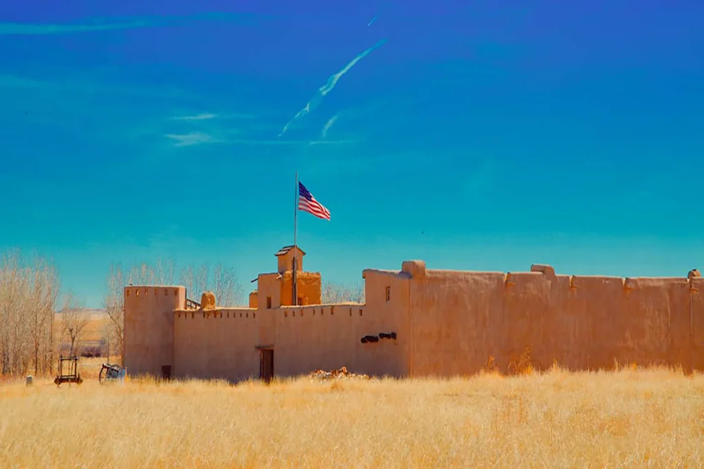 A historic adobe fort with an American flag on top, surrounded by dry grass under a clear blue sky.