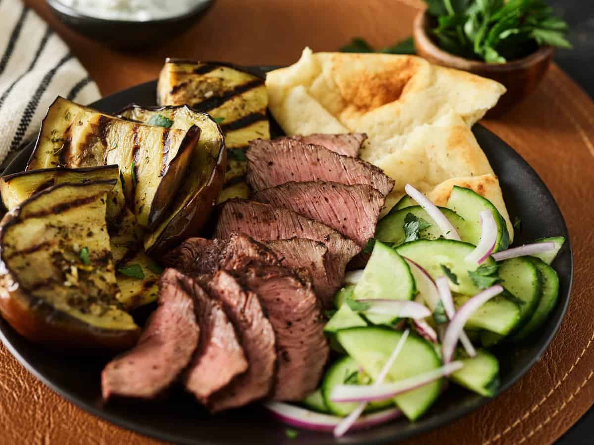 A plate with grilled eggplant, sliced steak, pita bread, cucumber slices with onions, and parsley garnish.