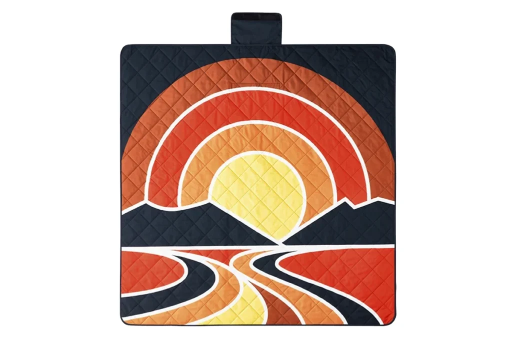 A quilted picnic blanket featuring a stylized sunset or sunrise design in red, orange, yellow, and black with mountains and a river.