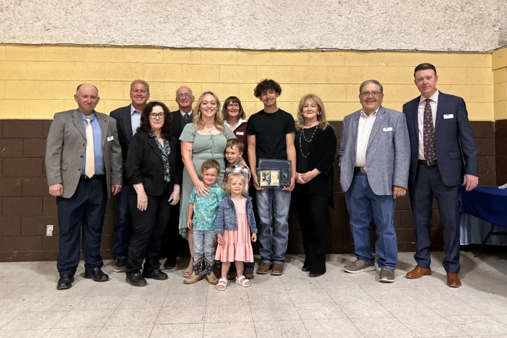 A group of people, including adults and children, pose indoors for a photo. One person in the center holds a framed award. Everyone is dressed in semi-formal attire.