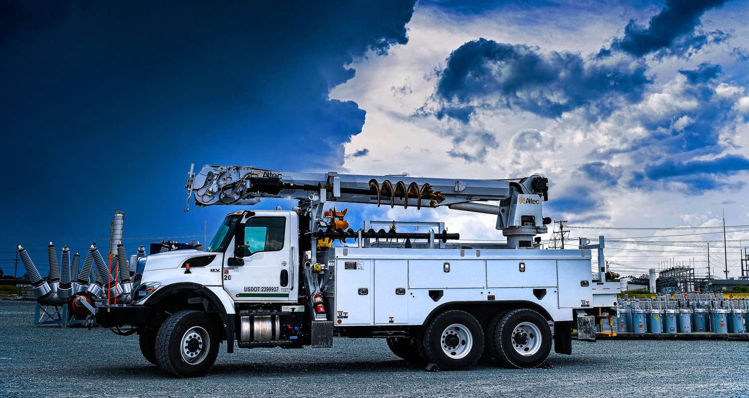 A white utility truck with an extended crane arm is parked on gravel, surrounded by electrical equipment under a partly cloudy sky.