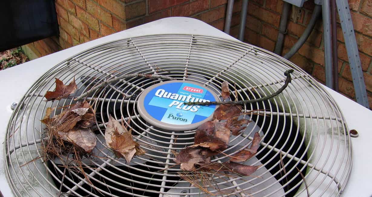 Air conditioning unit with leaves on the fan grille, positioned next to a brick wall.
