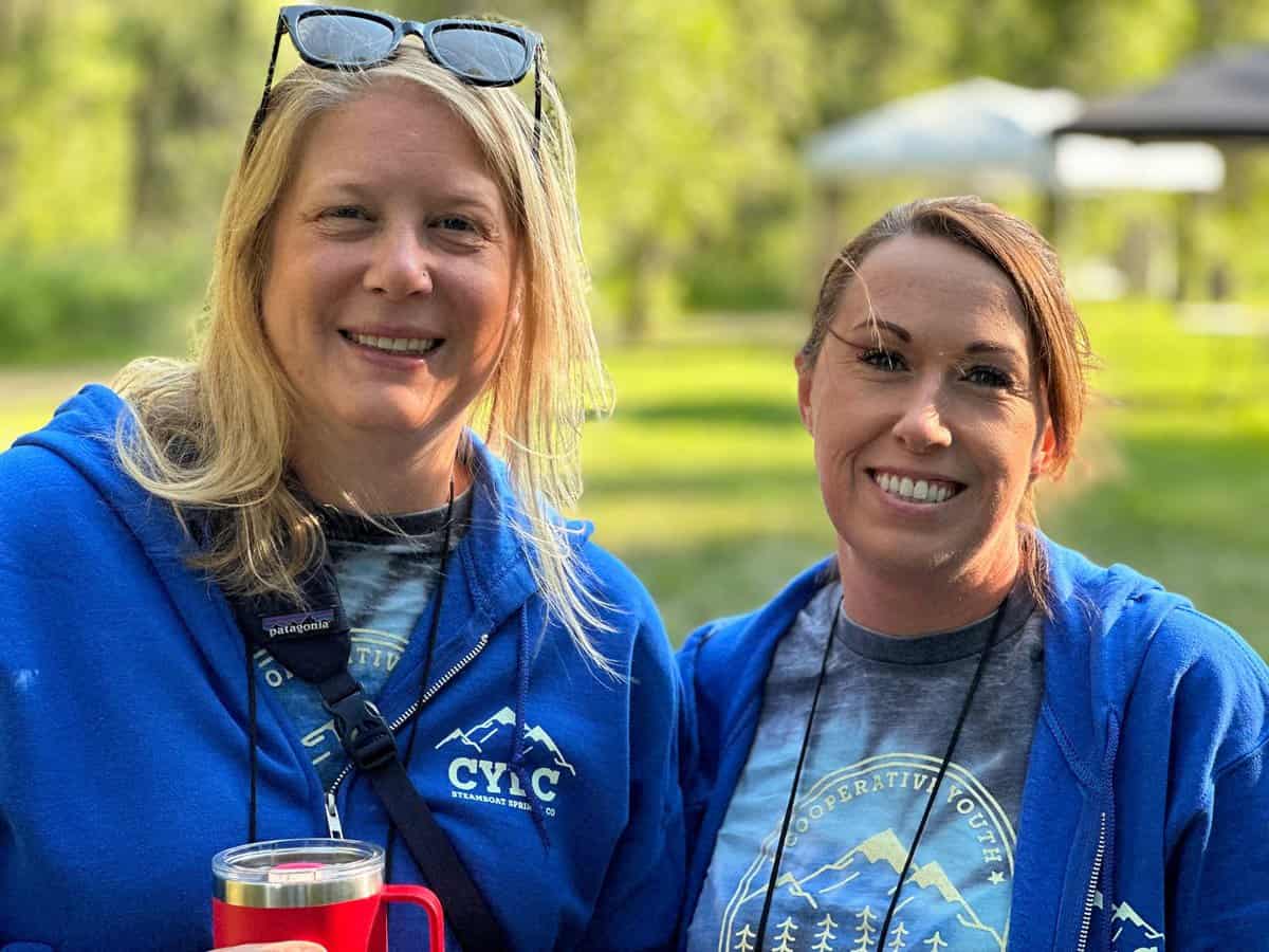 Two people outdoors wearing blue jackets with "CYLC" logos, smiling at the camera. One holds a red cup. Greenery and a structure are in the background.
