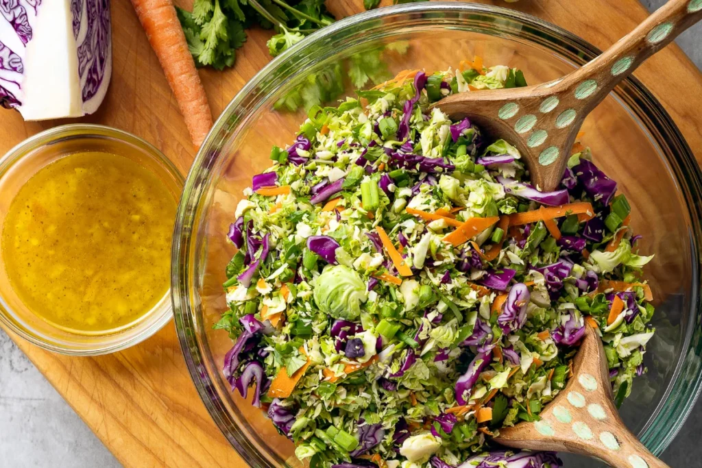 A glass bowl filled with chopped salad including cabbage, carrots, and greens, with two wooden serving spoons; a small bowl of salad dressing sits nearby.