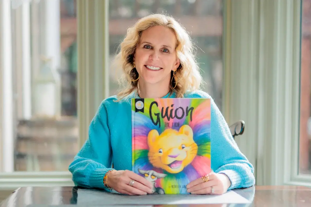 A person with wavy blonde hair holds a colorful book titled "Guión" with a smiling cartoon lion on the cover. They are sitting at a table in front of large windows.