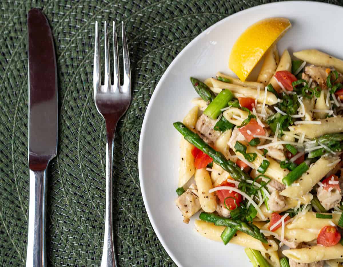 Plate of penne pasta with asparagus, tomatoes, cheese, and herbs next to a lemon slice. Fork and knife on a green woven placemat.
