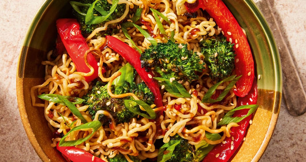 A bowl of noodles with broccoli, red bell peppers, and green onions, sprinkled with sesame seeds.