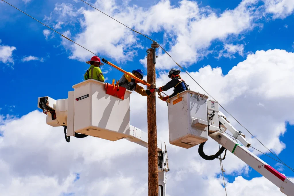Two utility workers in bucket trucks repair power lines on a wooden pole under a partly cloudy sky.