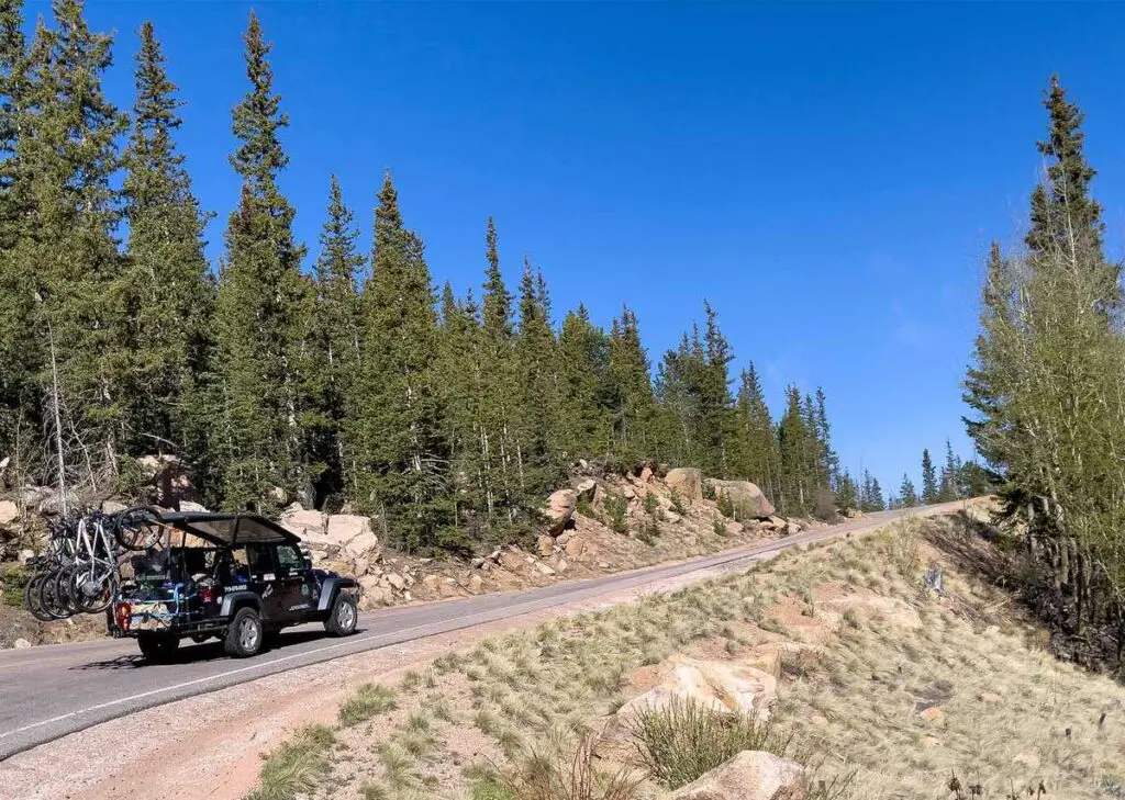 Bicycling: Get Out and Explore Truck with bicycles drives on a mountain road lined with pine trees under a clear blue sky.