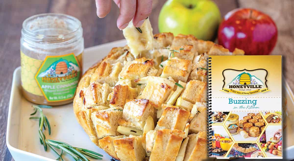 Pull-apart bread with rosemary and cheese on a plate, next to a jar of honey, apples, and a Honeyville cookbook. A hand is pulling a piece from the bread.