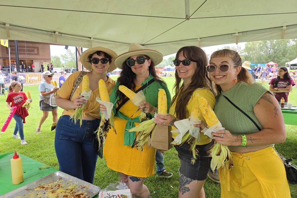 Four women standing under a tent at an outdoor event, each holding an ear of corn on the cob, smiling at the camera.