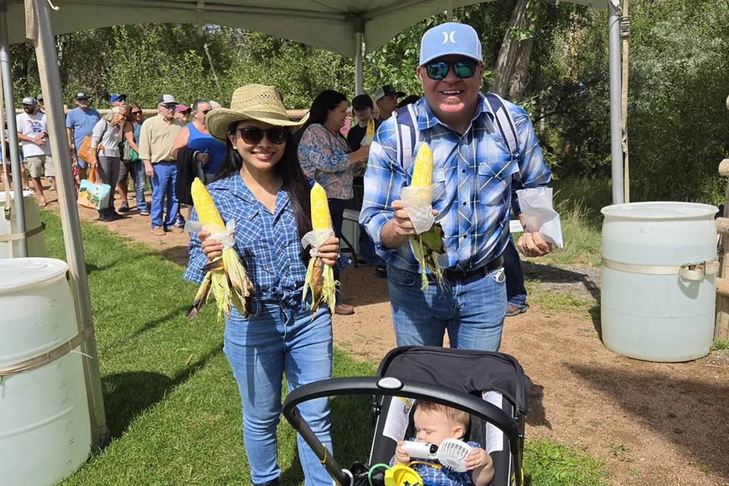 Two adults holding corn on the cob stand under a tent at an outdoor event, with a baby in a stroller in front of them and a group of people lined up in the background.