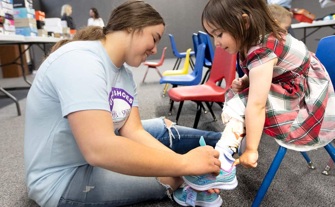 A woman fitting shoes on a young girl's foot. They are seated on the floor in a room with colorful chairs and tables filled with shoeboxes in the background.
