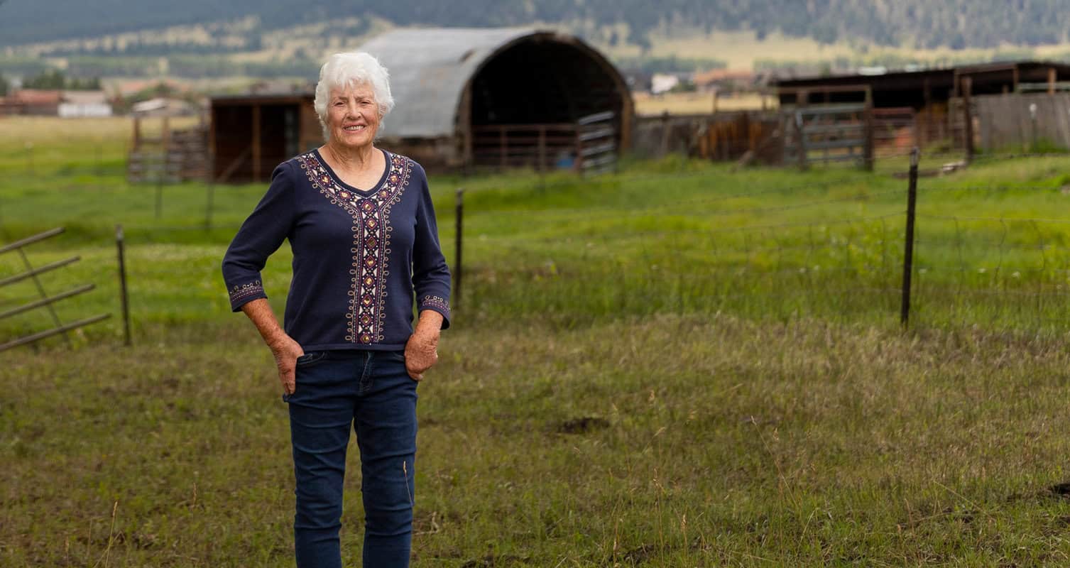 Elderly woman with white hair stands in a grassy field, wearing a blue top and jeans. Barns and hills are visible in the background.