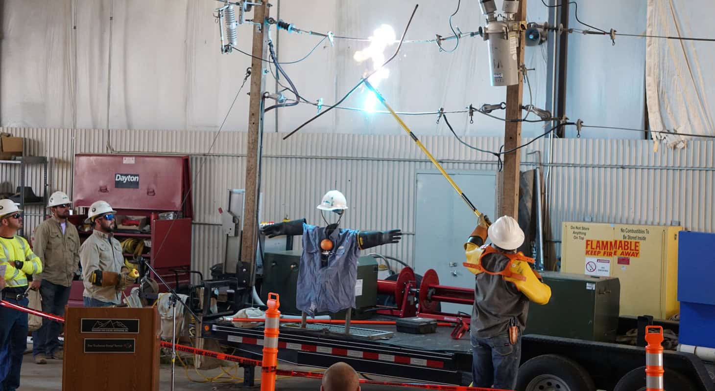 Workers demonstrate electrical safety precautions using a mannequin and high voltage equipment indoors.