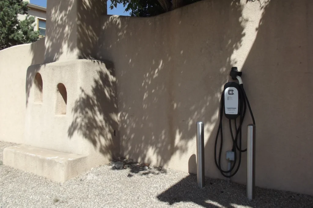 An electric vehicle charging station is mounted on an exterior stucco wall, with two metal posts in front and a bench built into the wall nearby.