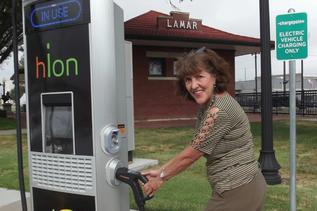 A woman charges an electric vehicle at a charging station near a building labeled "Lamar" and a sign indicating electric vehicle charging only.