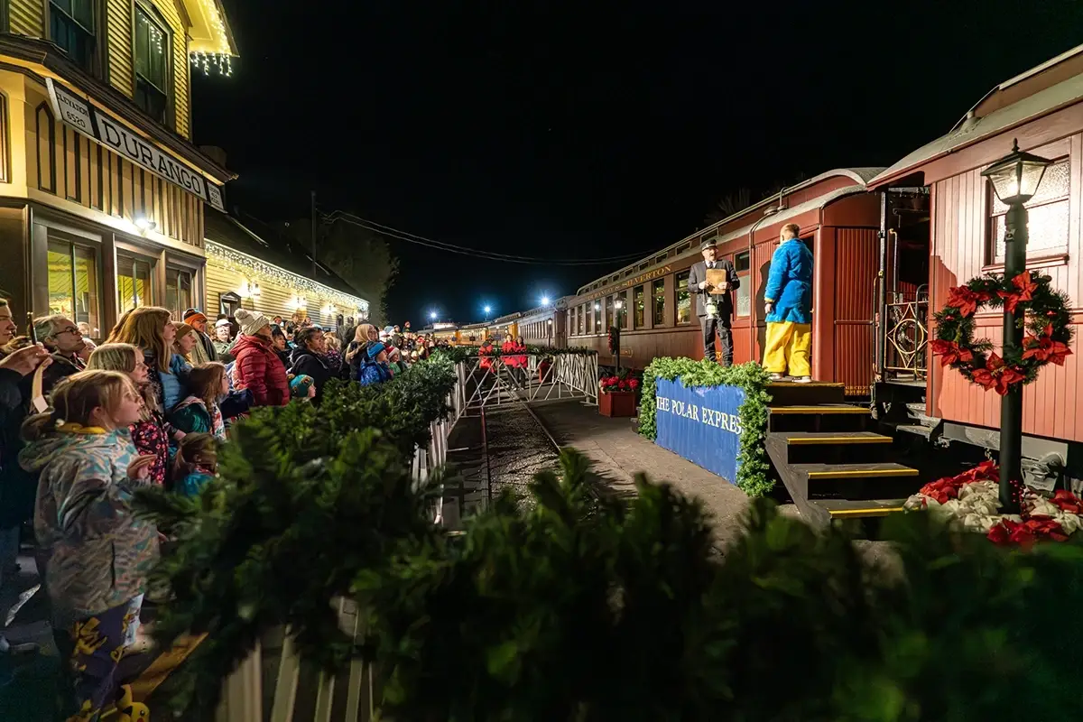 A crowd gathers at a festively decorated train station at night as performers stand on a platform beside a red train car labeled "The Polar Express.