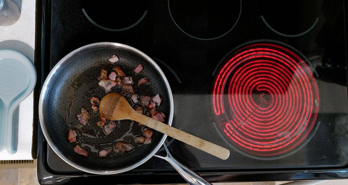 A frying pan with pieces of food being cooked on an electric stove, with one burner glowing red and a wooden spoon placed inside the pan.