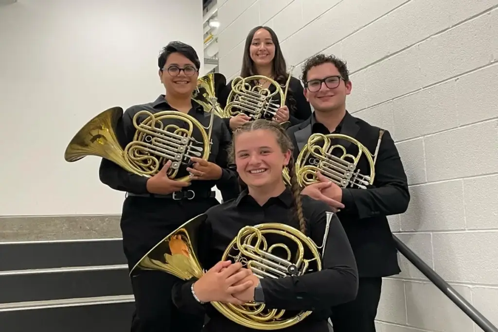 Four people in black attire stand on stairs holding French horns and smiling at the camera.