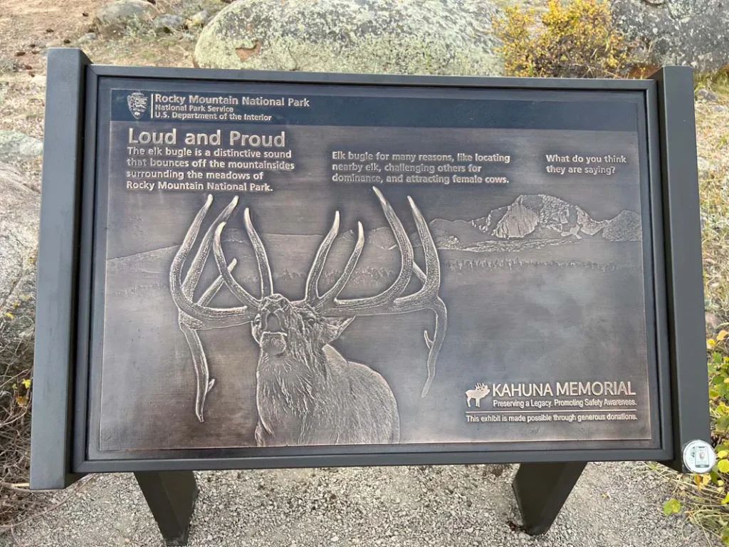 Informational sign at Rocky Mountain National Park with a raised elk illustration, describing elk bugling and its significance, mounted on a black metal stand outdoors.