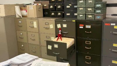A room filled with stacked filing cabinets and various boxes. An open drawer reveals papers and a small doll placed inside. Papers are scattered across a table in the foreground.