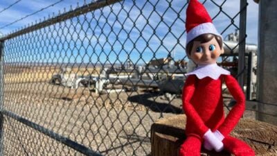 Elf on the Shelf doll in a red outfit sitting on a wooden post in front of a chain-link fence with industrial equipment in the background under a blue sky.
