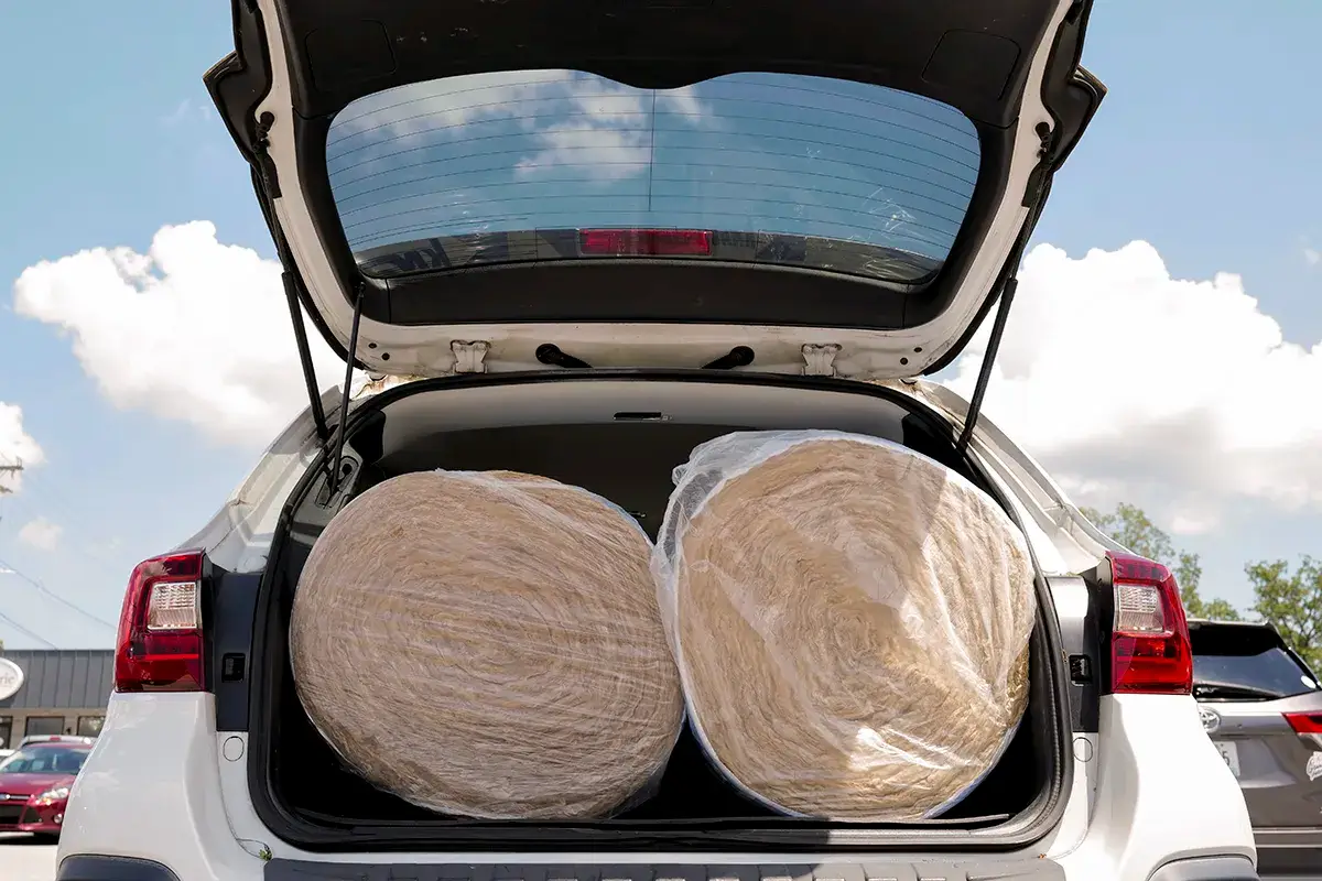 Two large rolls of insulation wrapped in plastic are loaded in the open trunk of a white SUV, with the sky and clouds visible through the rear window.