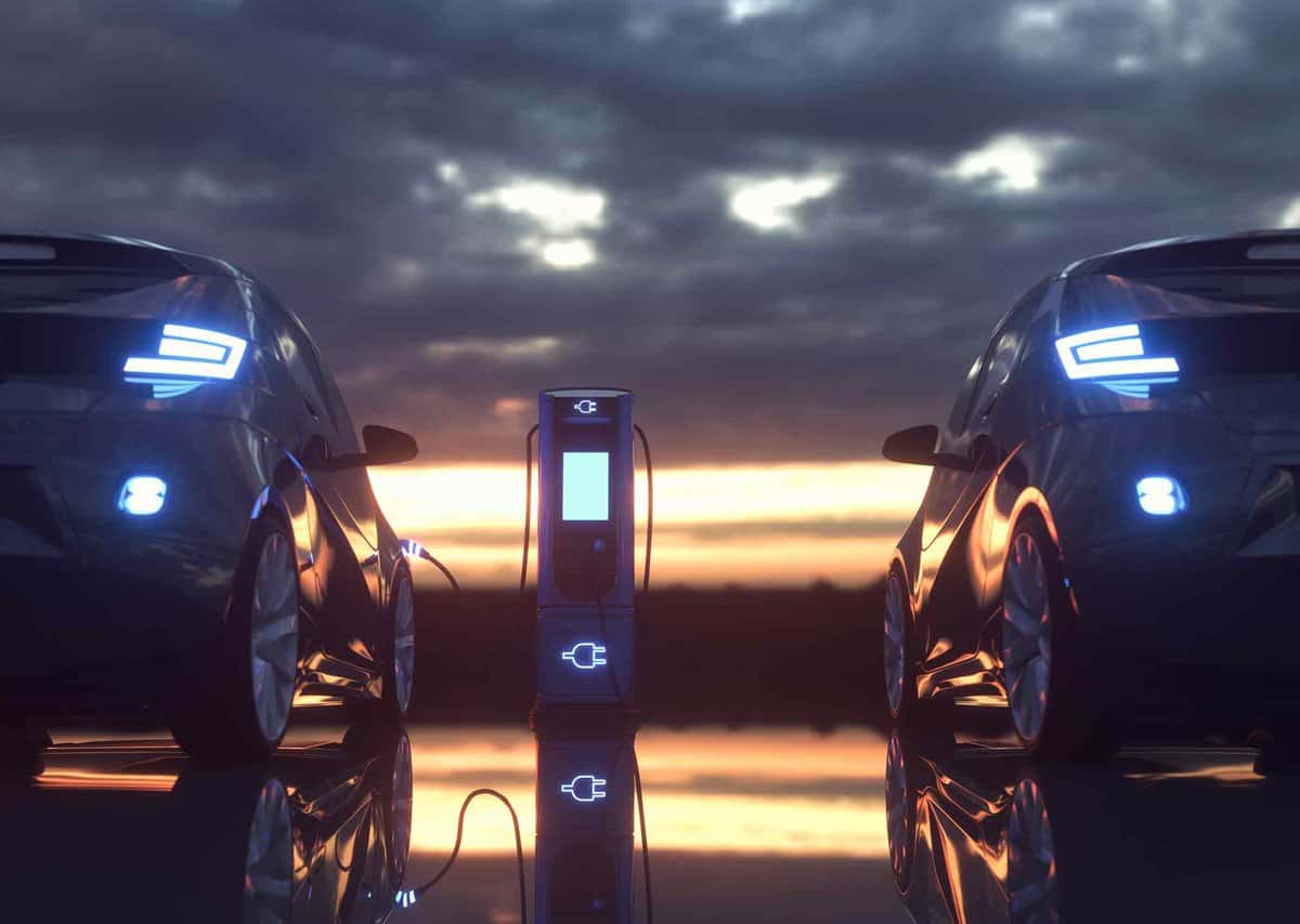 Two electric cars parked on either side of a charging station at sunset, reflecting on a glossy surface under a cloudy sky.