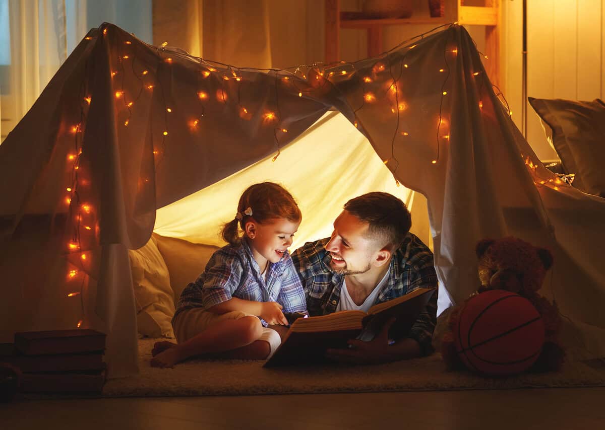 A father and daughter read a book inside a homemade tent, illuminated by string lights. A teddy bear and a basketball are nearby.