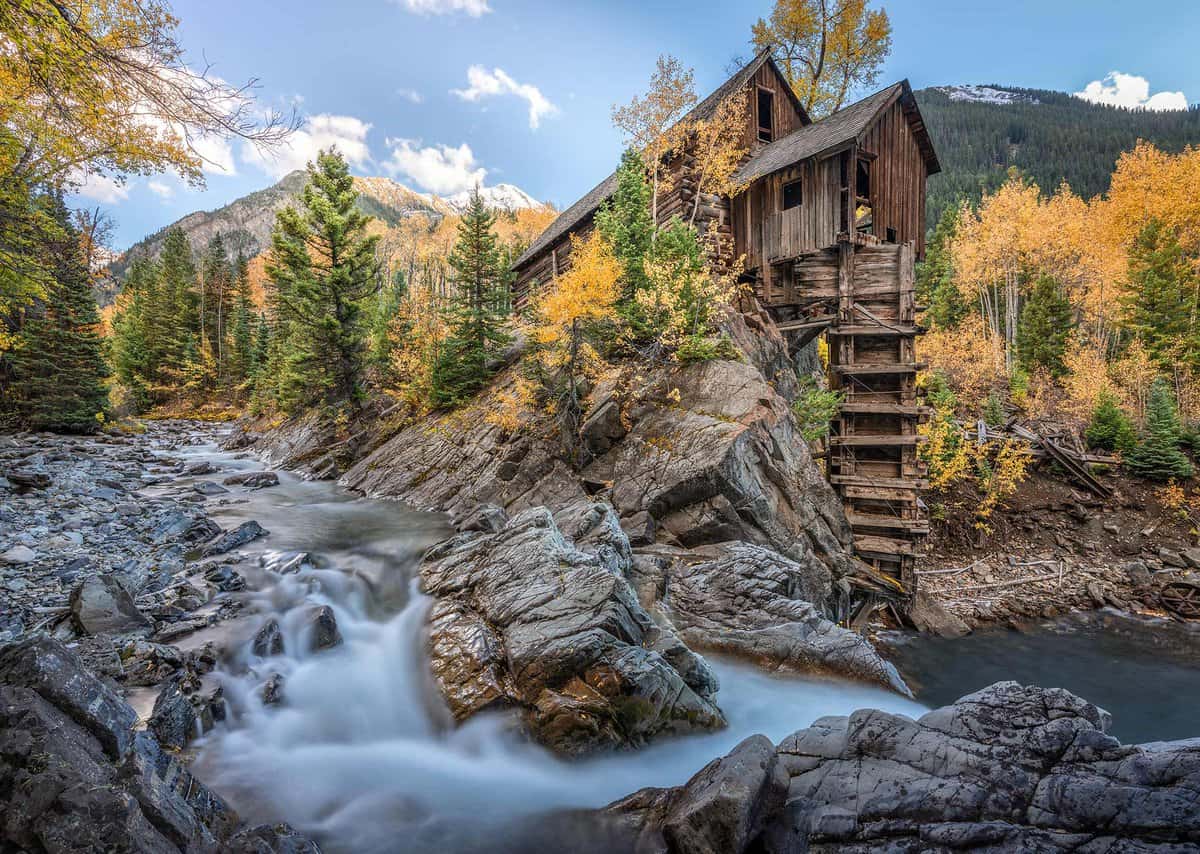 A rustic wooden cabin stands on a rocky cliff beside a flowing stream, surrounded by lush autumn trees and mountain scenery under a clear sky.