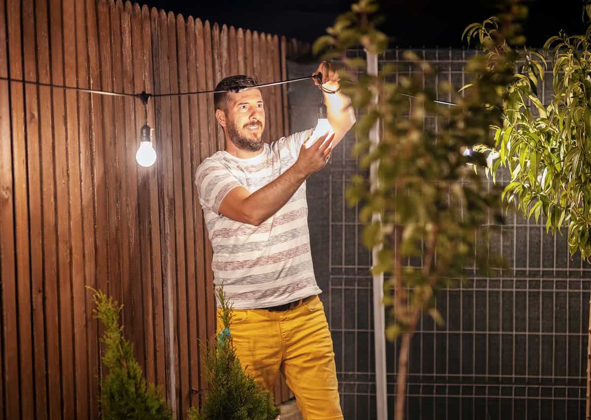 Man in striped shirt and yellow pants adjusts string lights in a garden with wooden fencing and greenery.