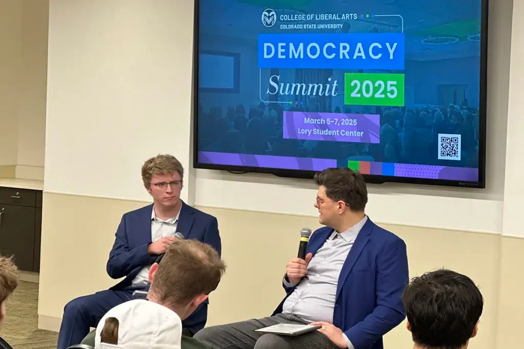 Two men in suits sit and talk on stage; one holds a microphone. Behind them is a screen displaying "Democracy Summit 2025" at Colorado State University.