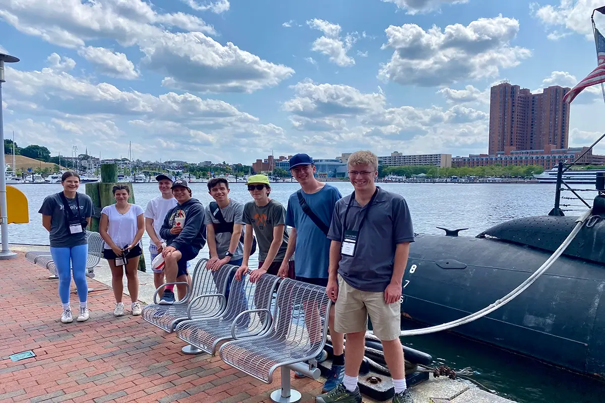 A group of eight people stands and sits by a waterfront next to a submarine, with city buildings and a partly cloudy sky in the background.