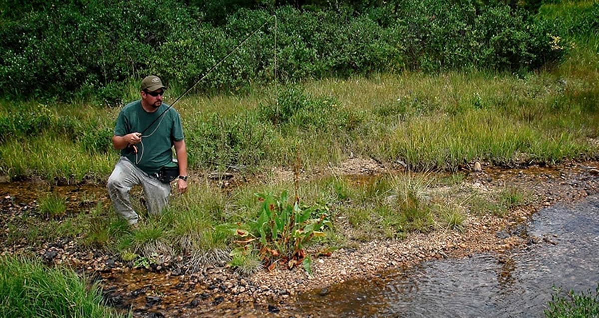 A man wearing outdoor gear kneels by the edge of a small stream while holding a fishing rod. Grass and shrubs are in the background.