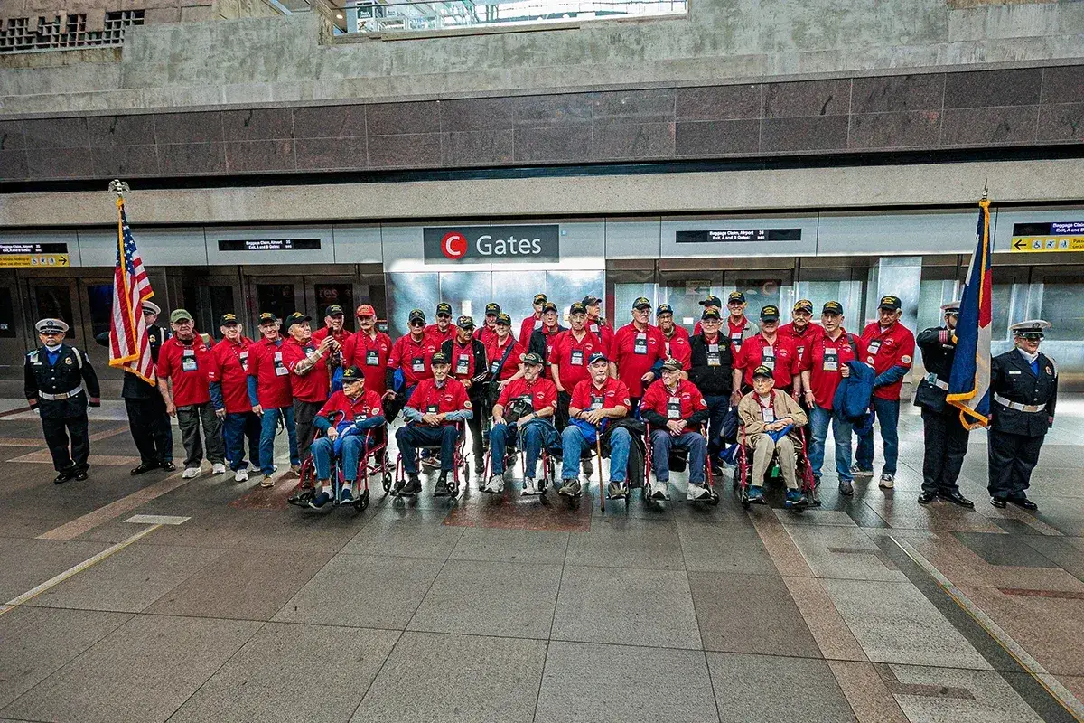 A group of veterans and volunteers in red shirts pose for a photo at an airport in front of signs for C Gates, with two uniformed individuals holding flags at each end.