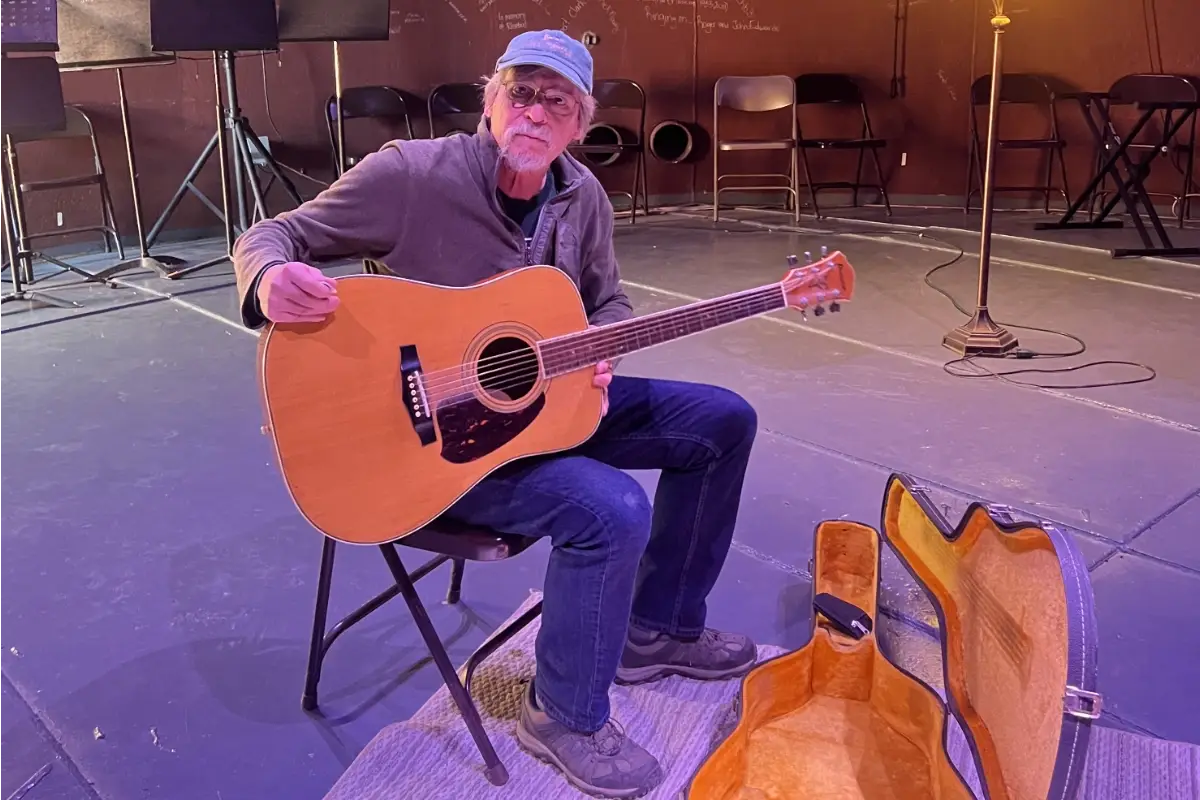 An older man wearing glasses, a cap, and casual clothes sits on a chair on stage, holding an acoustic guitar. An open guitar case is on the floor beside him. Several empty chairs and stands are in the background.