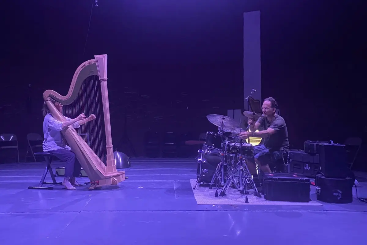 A person playing a large wooden harp faces another person playing a drum set on a dimly lit stage with a purple hue. Empty chairs are visible in the background.