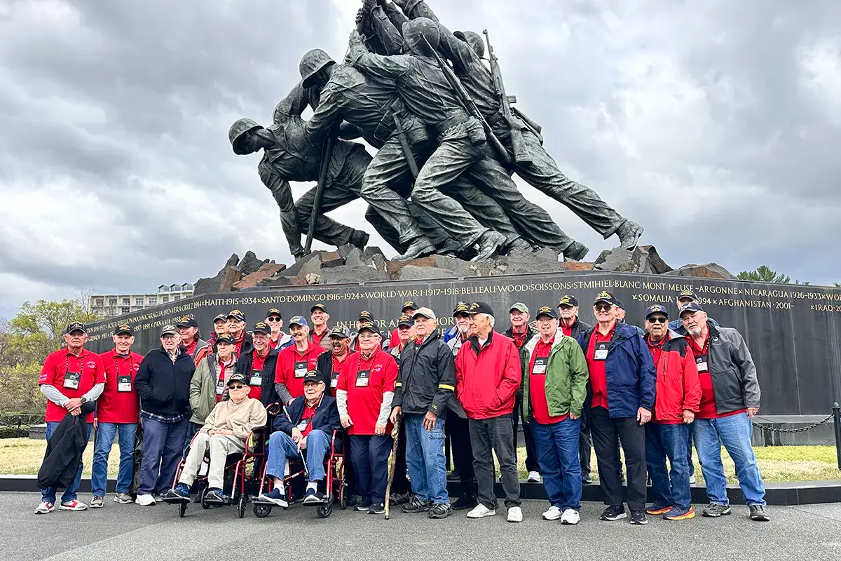 A group of people, some in red shirts and a few in wheelchairs, pose in front of the Marine Corps War Memorial statue depicting the flag raising at Iwo Jima.