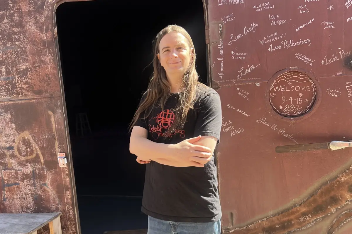 A person with long hair stands smiling with arms crossed in front of a large, rust-colored metal door covered in handwritten names and messages in white ink. Sunlight illuminates the scene.