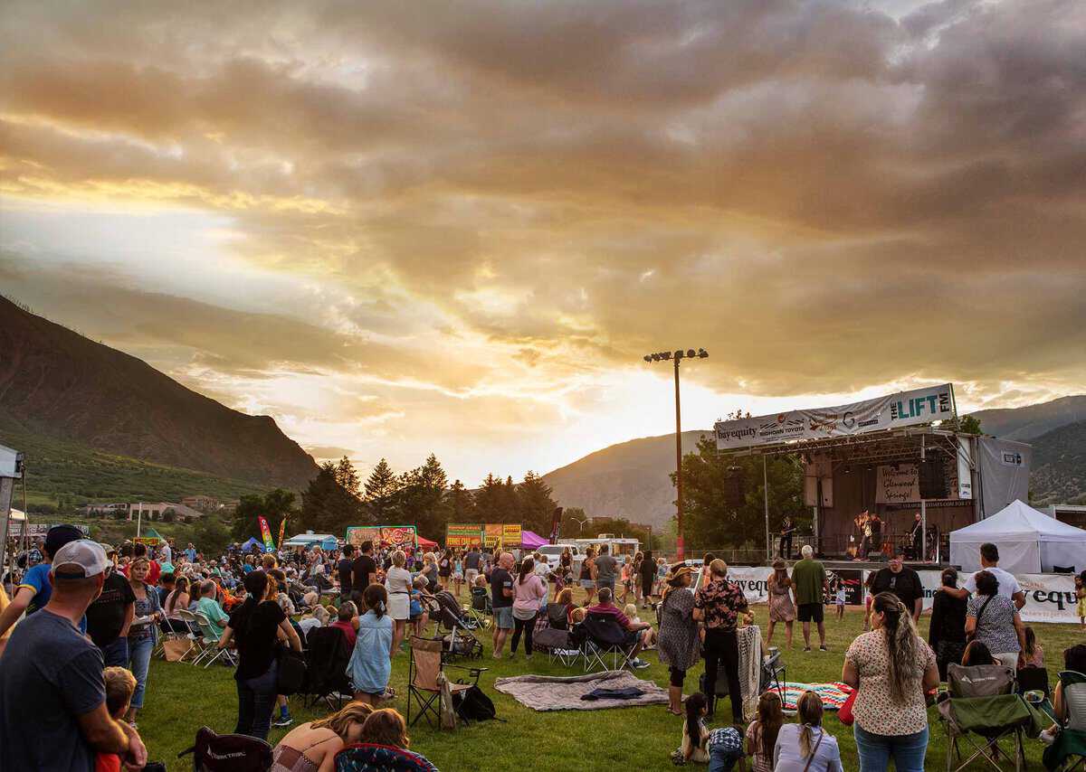 Outdoor concert at sunset with a crowd sitting and standing on a grassy field, a stage on the right, and mountains in the background under a cloudy sky.