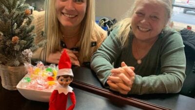 Two women smiling behind a counter with a bowl of candies. An Elf on the Shelf doll sits in front of them. A small decorated Christmas tree is on the counter.