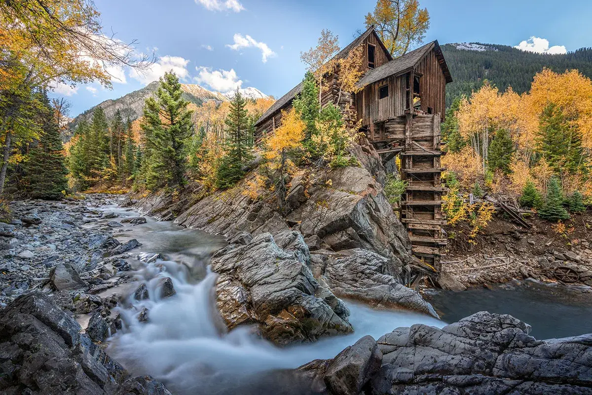 A wooden mill perched on a rocky slope beside a flowing river, surrounded by autumn trees and mountains in the background.
