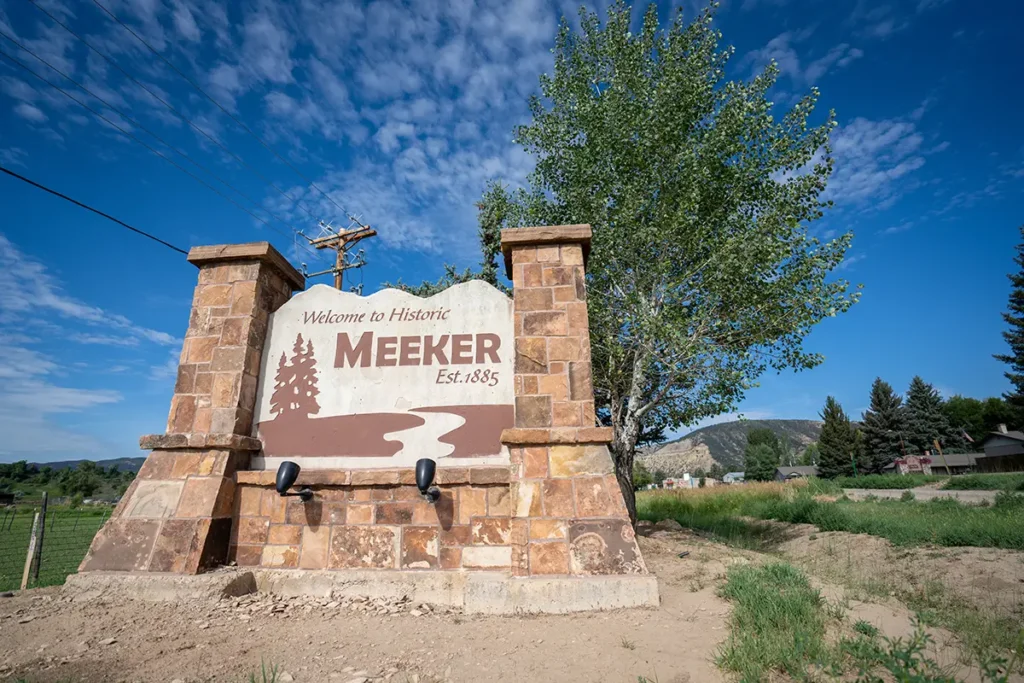 A large stone sign reads "Welcome to Historic Meeker Est. 1885" next to a tree and grassy area under a blue sky with scattered clouds.